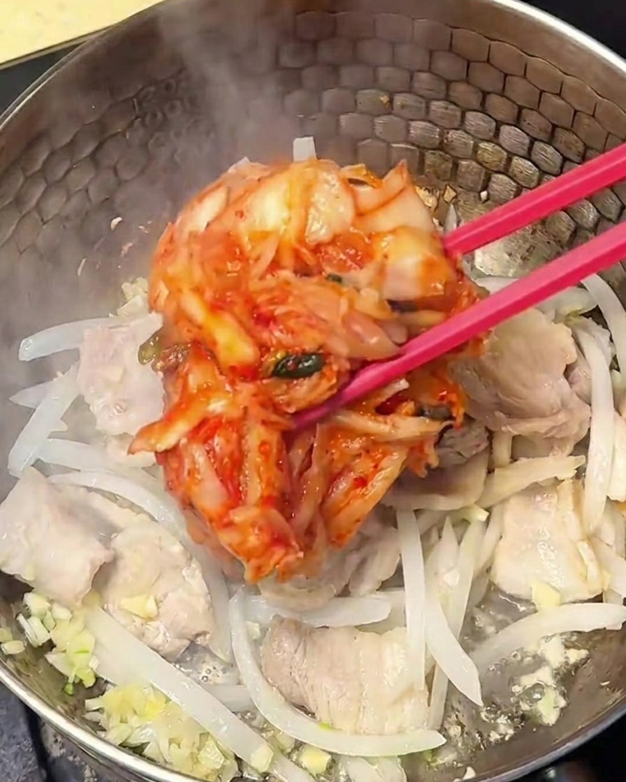 Kimchi rosso condito che viene mescolato con il composto di pancetta e cipolla nella pentola.