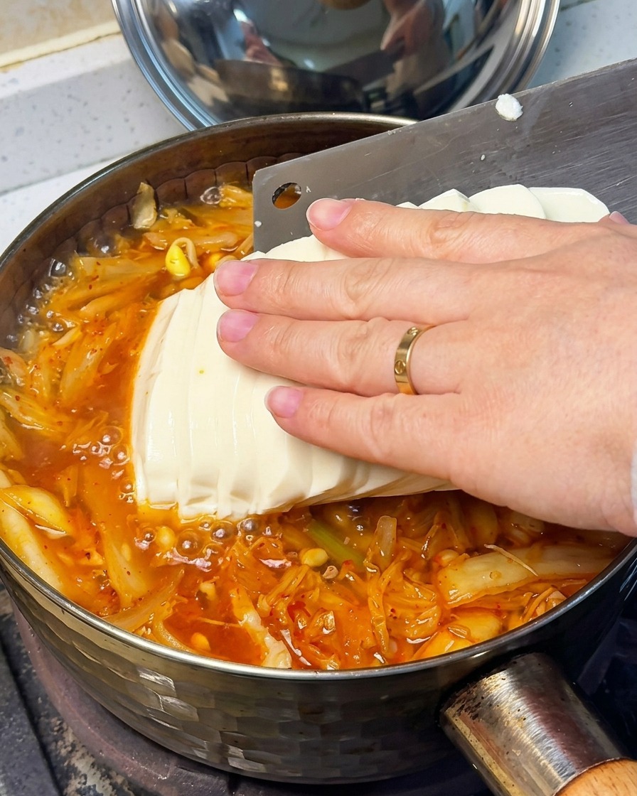 Primo piano di una mano che posiziona delicatamente fette di tofu vellutato nella zuppa di kimchi rossa.