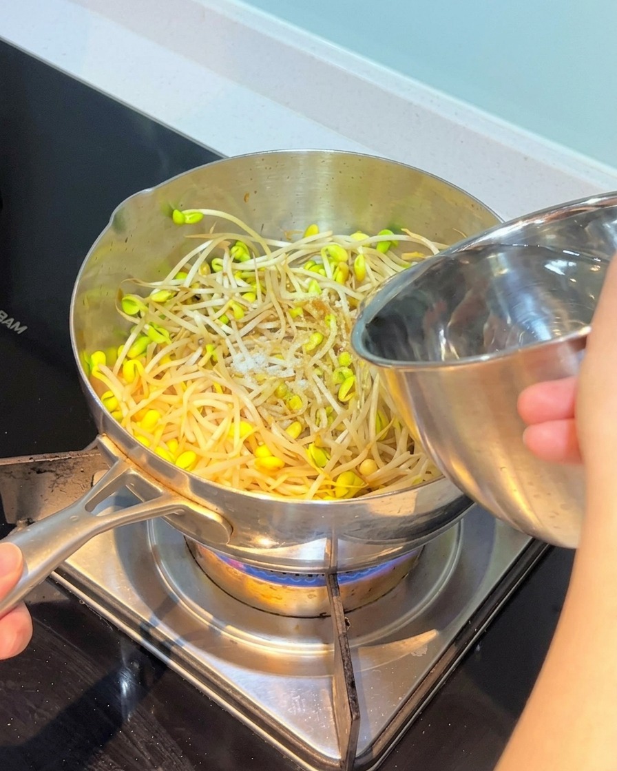 Acqua versata da una ciotola di acciaio inox in una pentola contenente germogli di soia e altri ingredienti saltati.