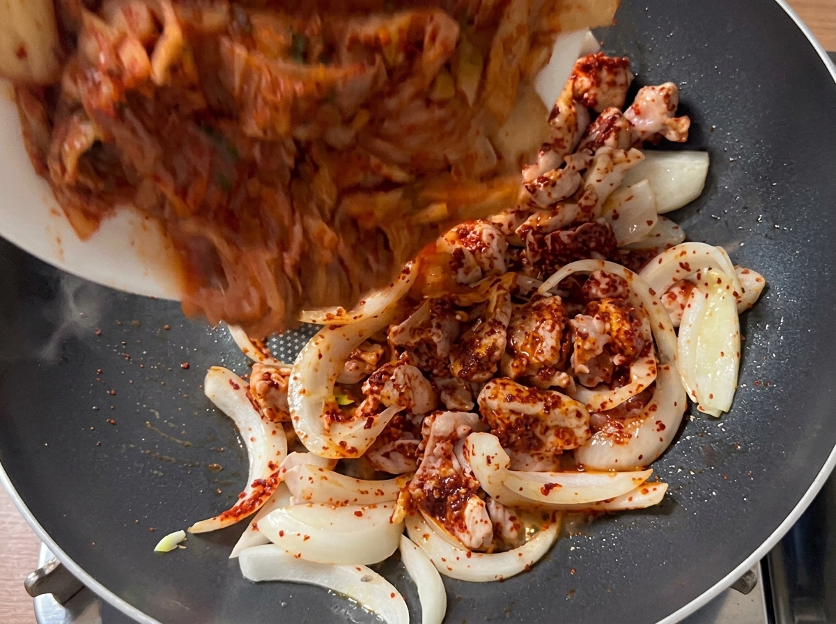 Pezzi di kimchi a misura di boccone versati nella padella con il mix di maiale e cipolla rosolato.