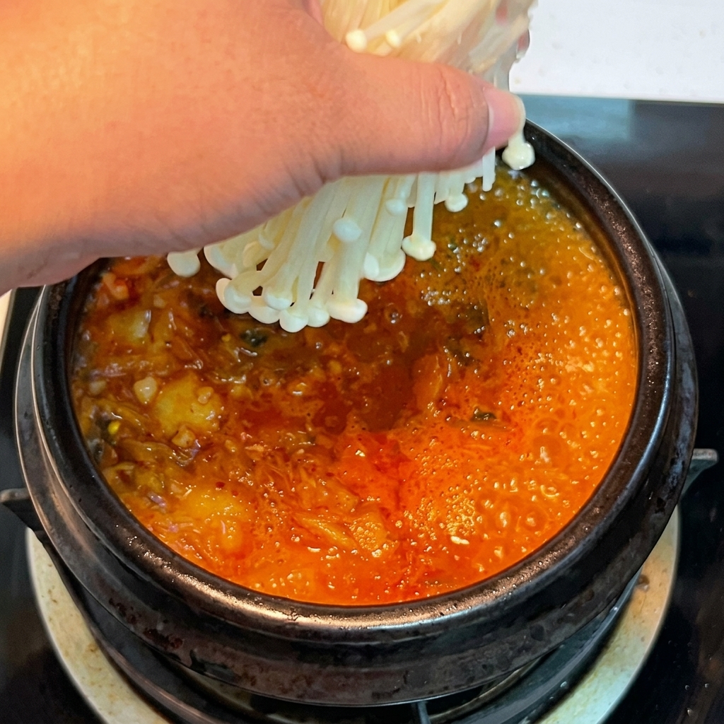 Una mano che mette un mazzetto di funghi enoki freschi nello stufato di kimchi rosso che ribolle.