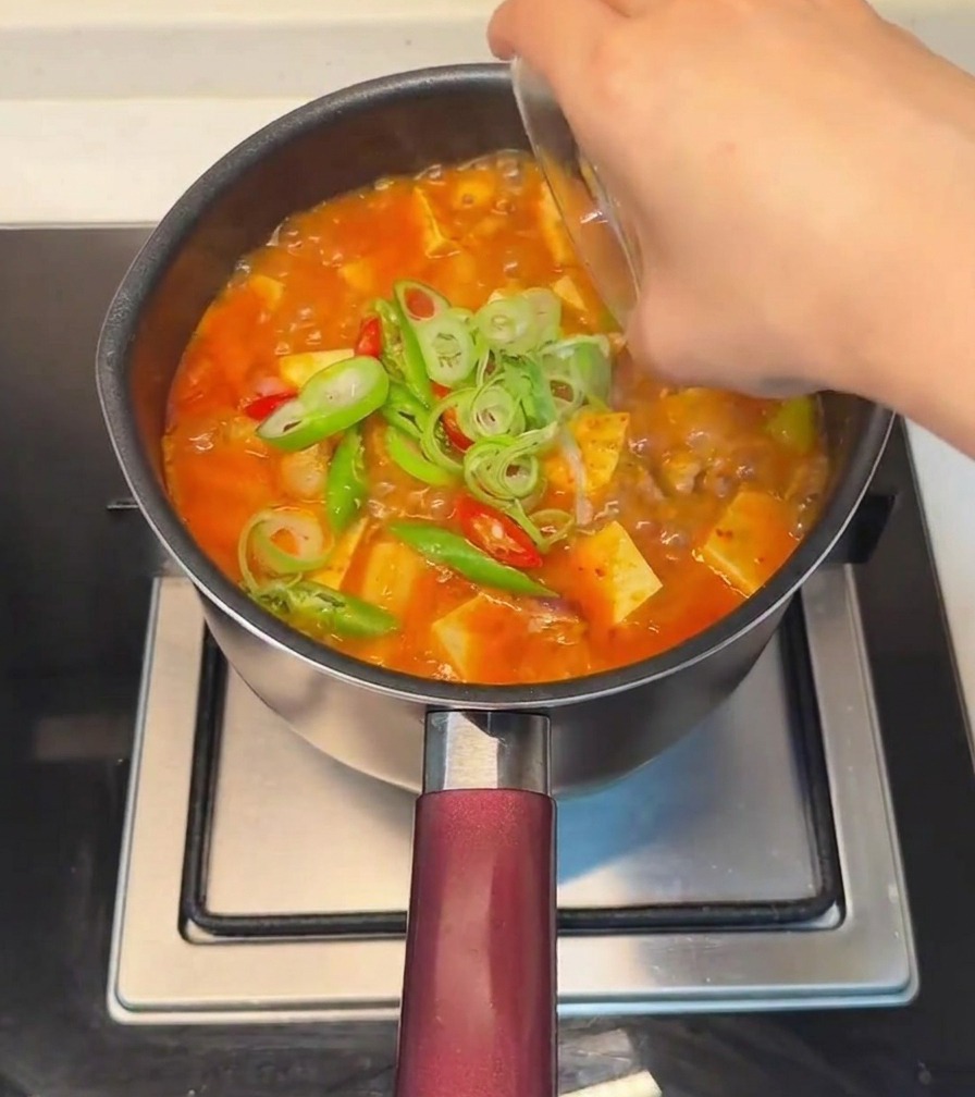 Una mano che cosparge cipollotti affettati e peperoncini rossi e verdi freschi sopra lo stufato di tofu finito.