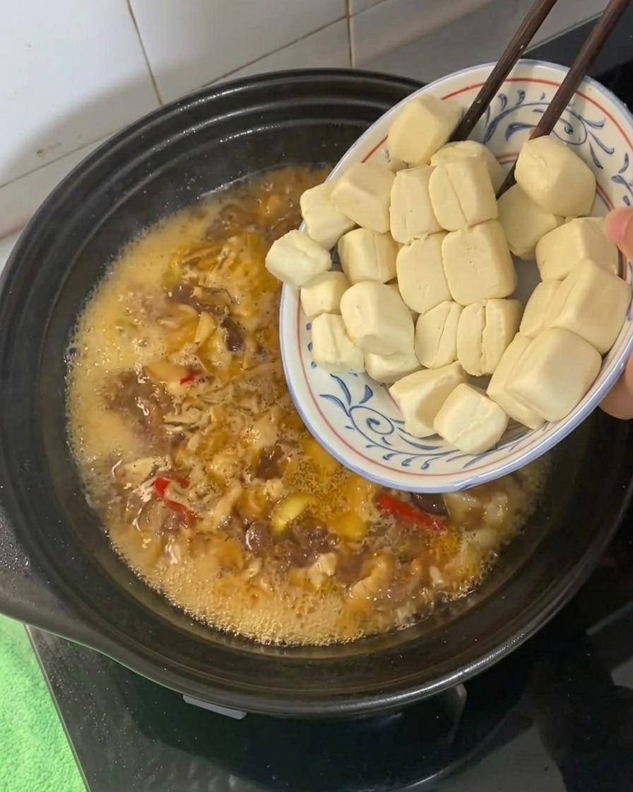 Piccoli cubetti di tofu aggiunti a un liquido di brasatura dorato che bolle in una pentola di coccio.