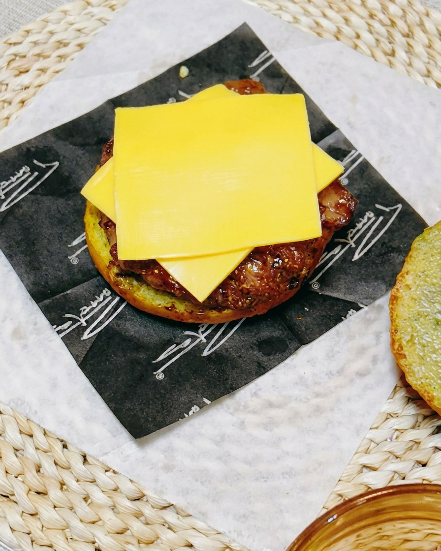 Un primo piano dellassemblaggio del burger che mostra una fetta di formaggio appoggiata sulla cotoletta di pollo, sormontata da rondelle di cetriolo fresco.