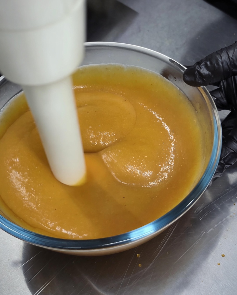 Un frullatore a immersione utilizzato per frullare una densa salsa dorata in una ciotola di vetro.