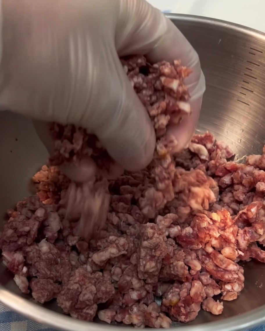 Una vista ravvicinata di una mano guantata che modella della carne di manzo macinata fresca in una pallina compatta.