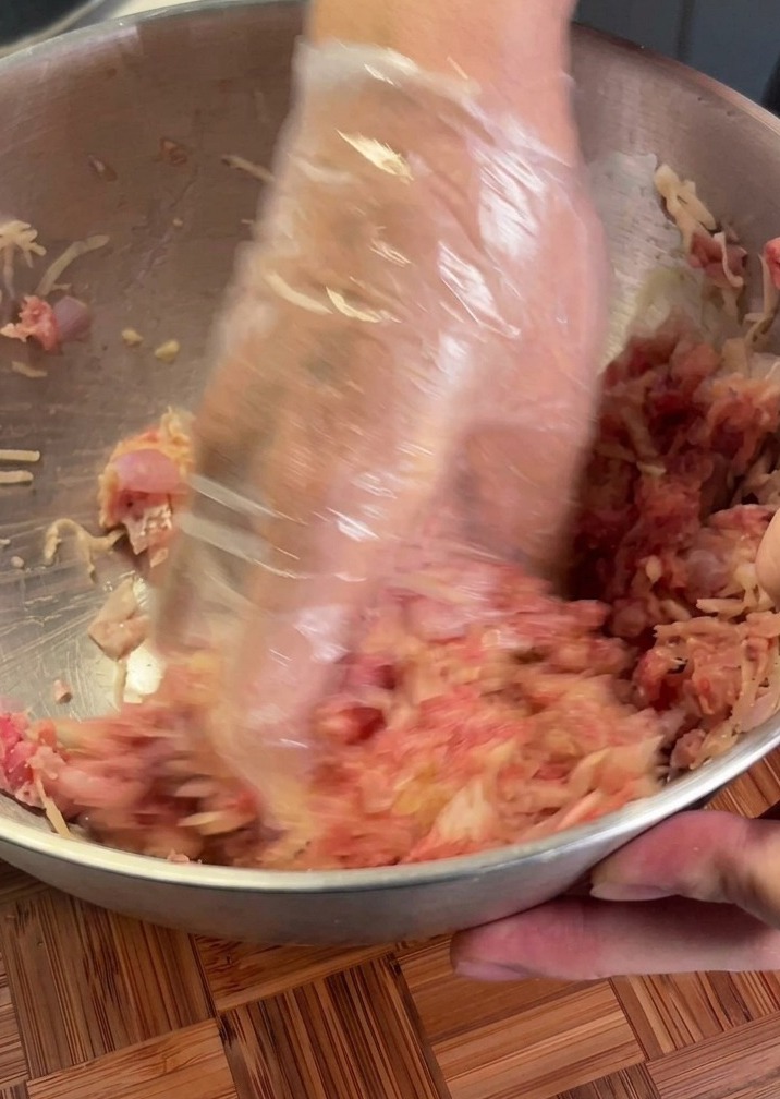 Mani con un guanto di plastica che mescolano manzo crudo e patata grattugiata in una ciotola di metallo.
