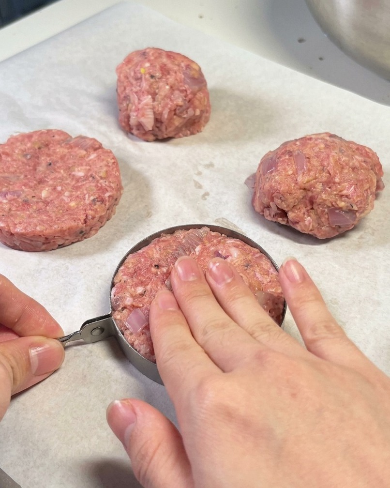 Mani che usano un coppa-pasta in metallo per modellare un hamburger su carta da forno.