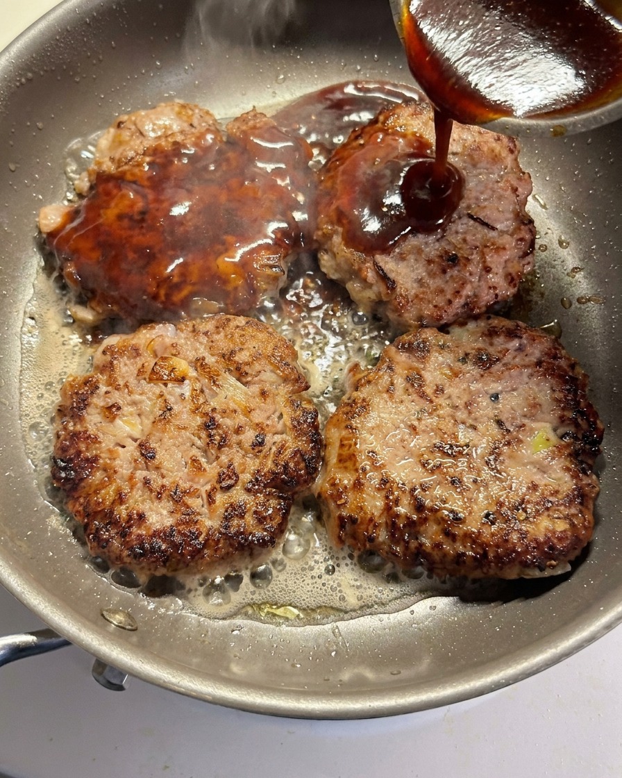 La salsa viene versata sopra quattro hamburger in doratura in una padella.
