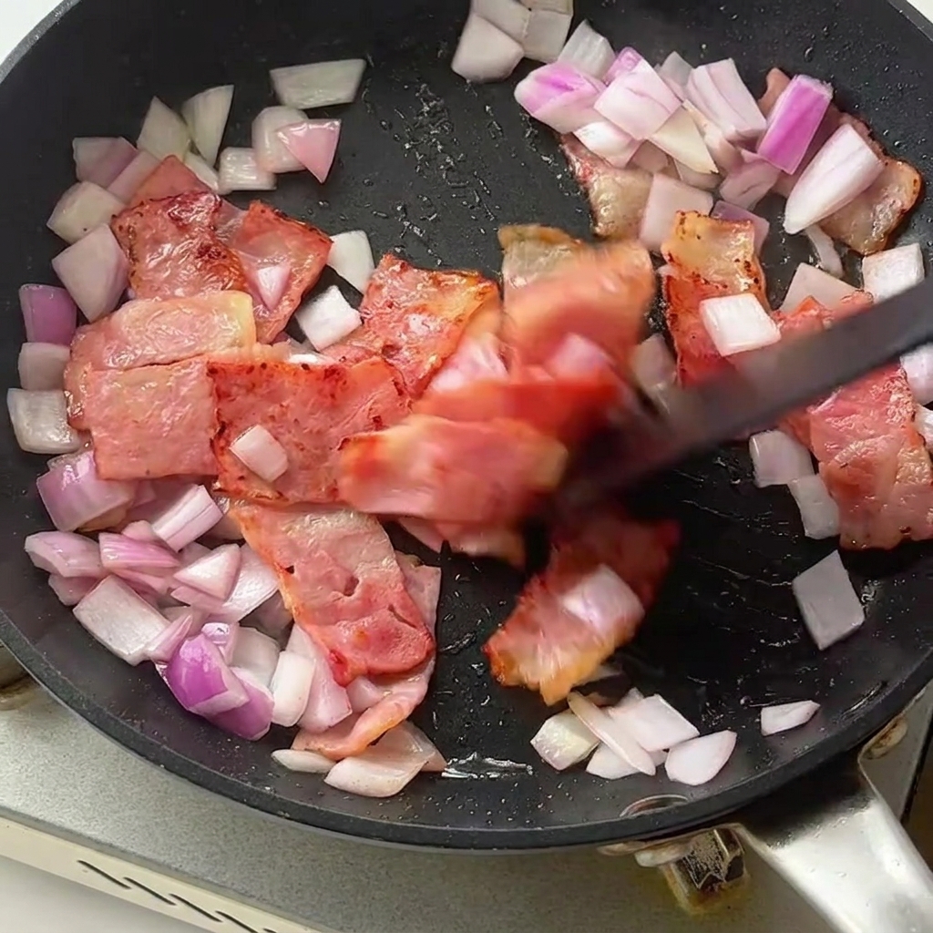 Fusilli e asparagi verdi inseriti nella padella con pancetta dorata e cipolle.