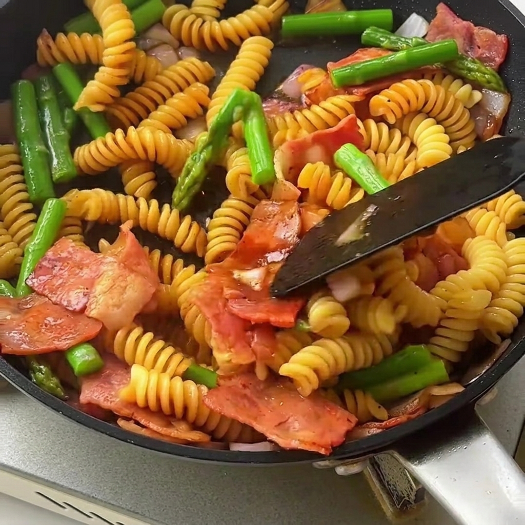 Una spatola che mescola i fusilli conditi, gli asparagi croccanti e la pancetta saporita in una grande padella.