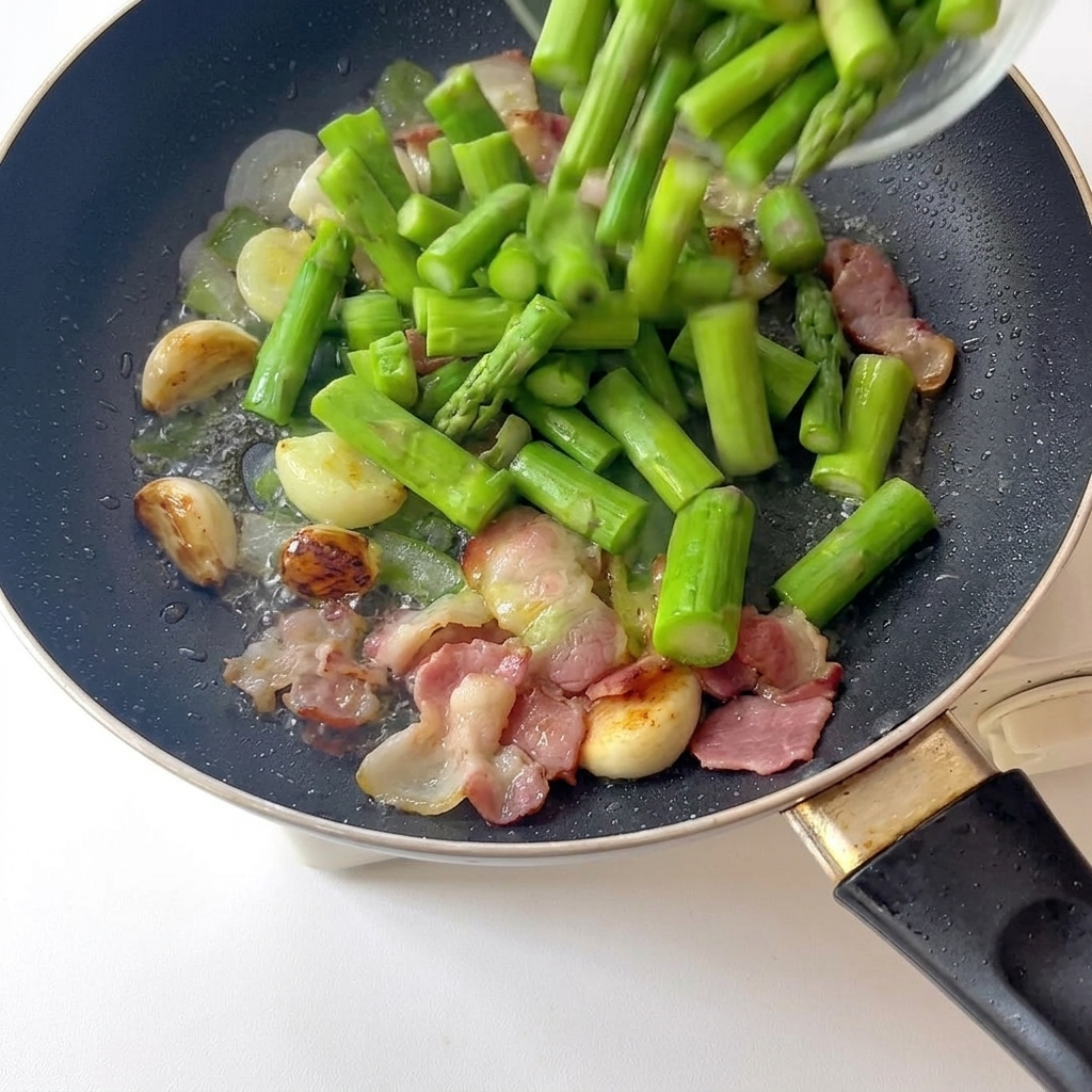 Pezzi di asparagi verde brillante appena sbollentati versati in una padella contenente aglio fritto e pancetta rosolata.