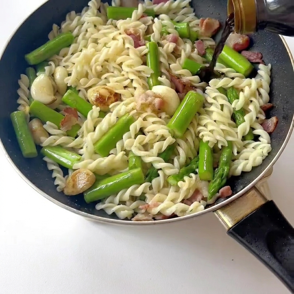 Primo piano di una spatola che mescola pasta, pancetta e asparagi in padella mentre viene versata la salsa di soia chiara.