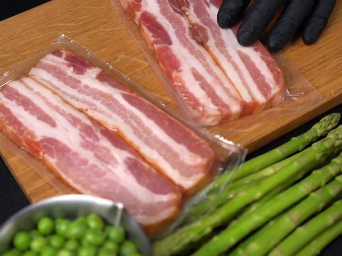 Primo piano di una mano con guanto che usa un coltello per tagliare strisce spesse di bacon crudo su un tagliere di legno.