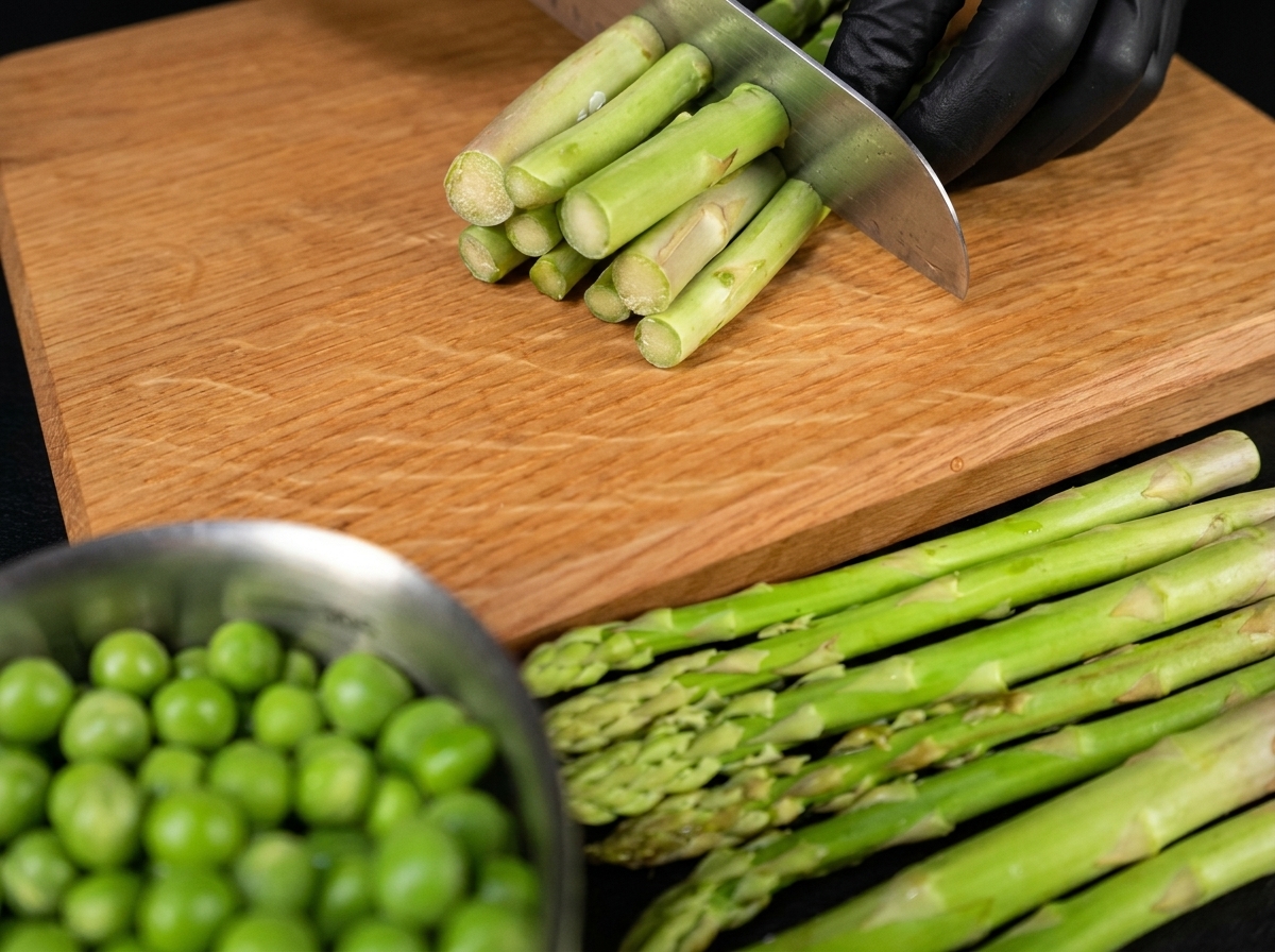 La mano di uno chef con un coltello che taglia gambi di asparagi verdi freschi in pezzi uniformi su una superficie di legno.