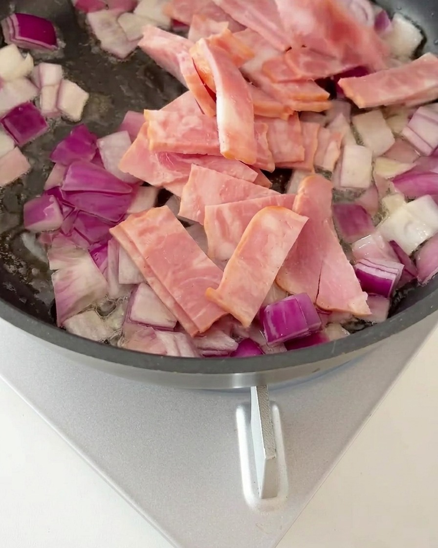 Pezzi di pancetta uniti alle cipolle soffritte in una padella, mentre iniziano a dorarsi.