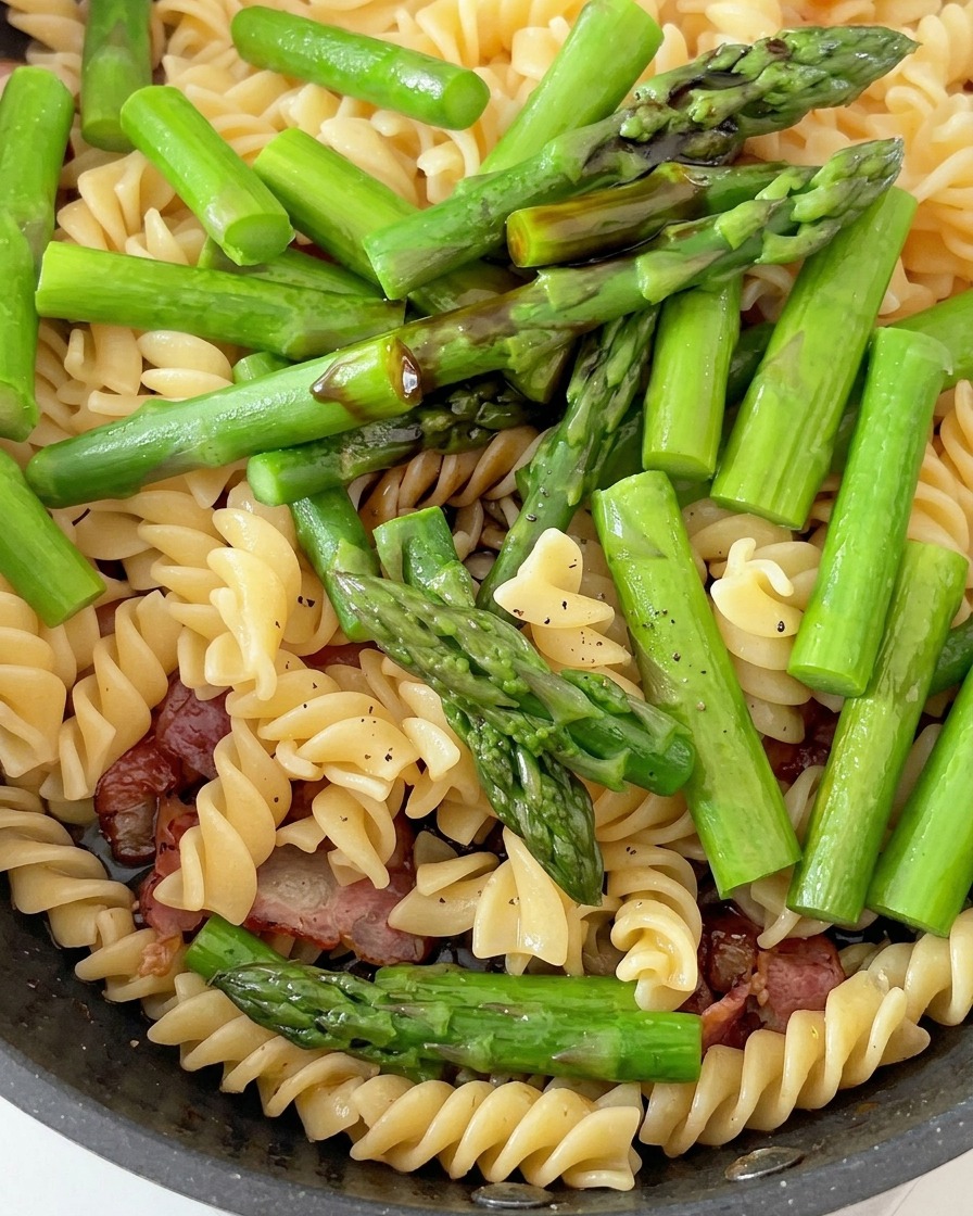 Una padella contenente pasta fusilli cotta, pezzi di asparagi verdi vivaci e pancetta cotta, condita con le salse.