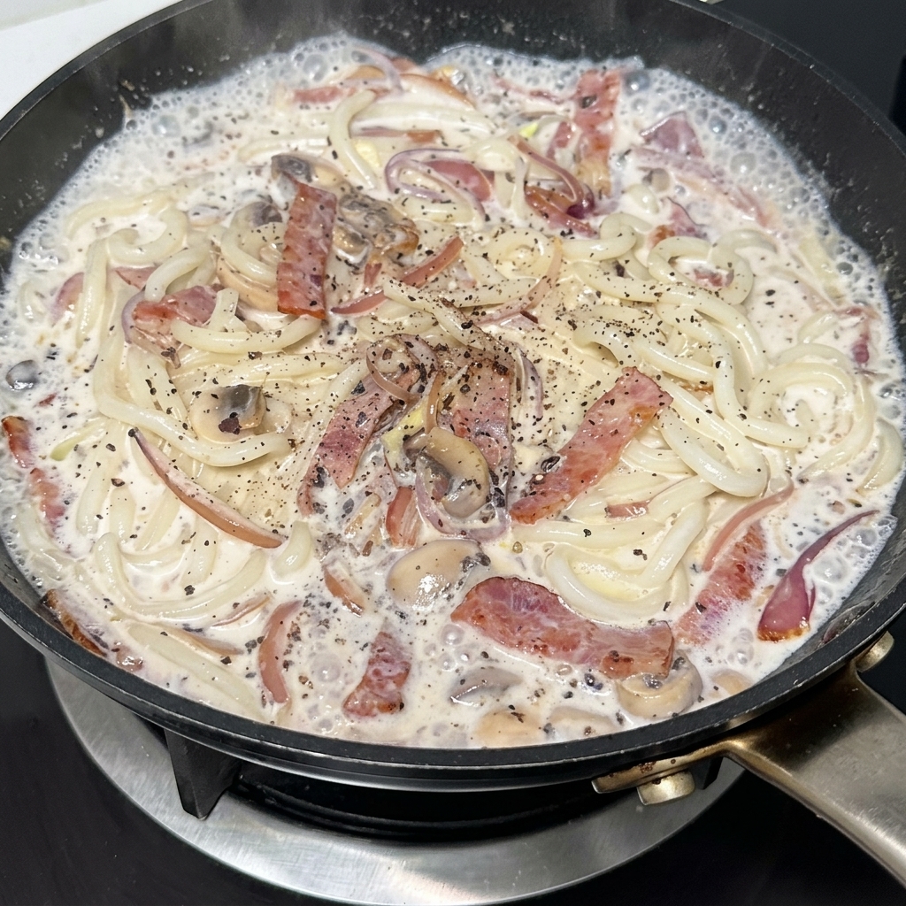 Una padella di udon cremosi con pancetta e funghi conditi con una spolverata di pepe nero.