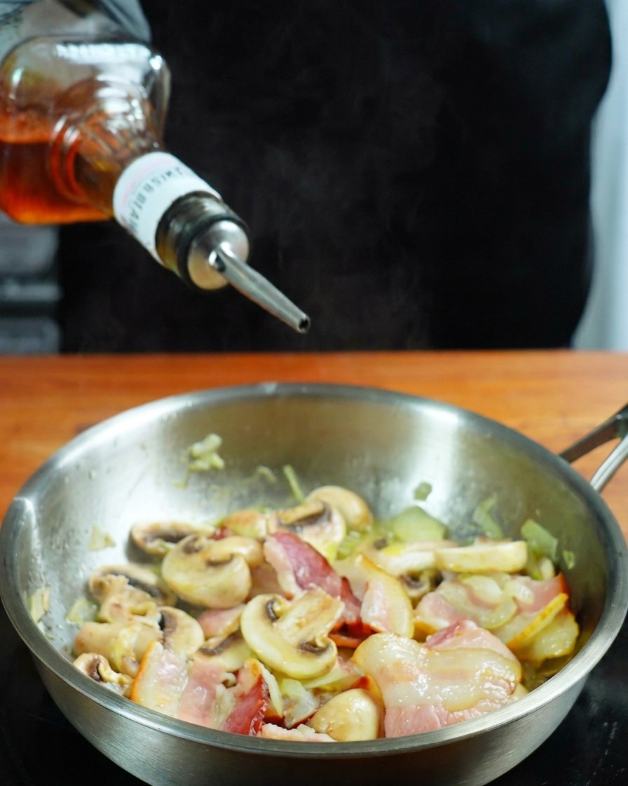 Whiskey versato da una bottiglia in una padella con pancetta e funghi.