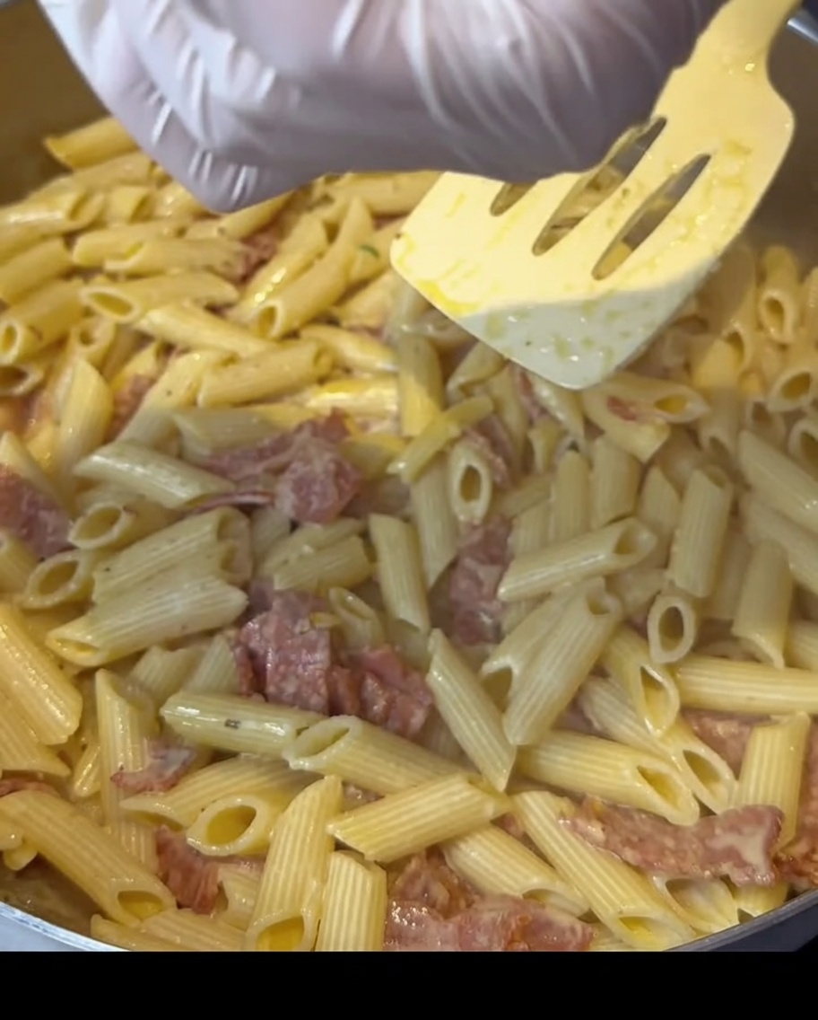 Mani con guanti che cospargono condimento su una padella di pasta cremosa al bacon.