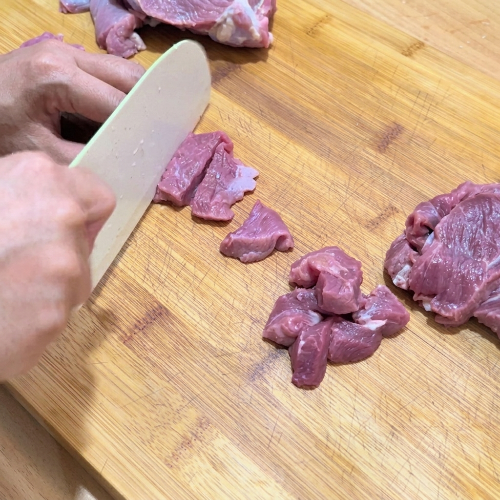 Mani che tagliano manzo crudo in cubetti piccoli e uniformi su un tagliere di legno.