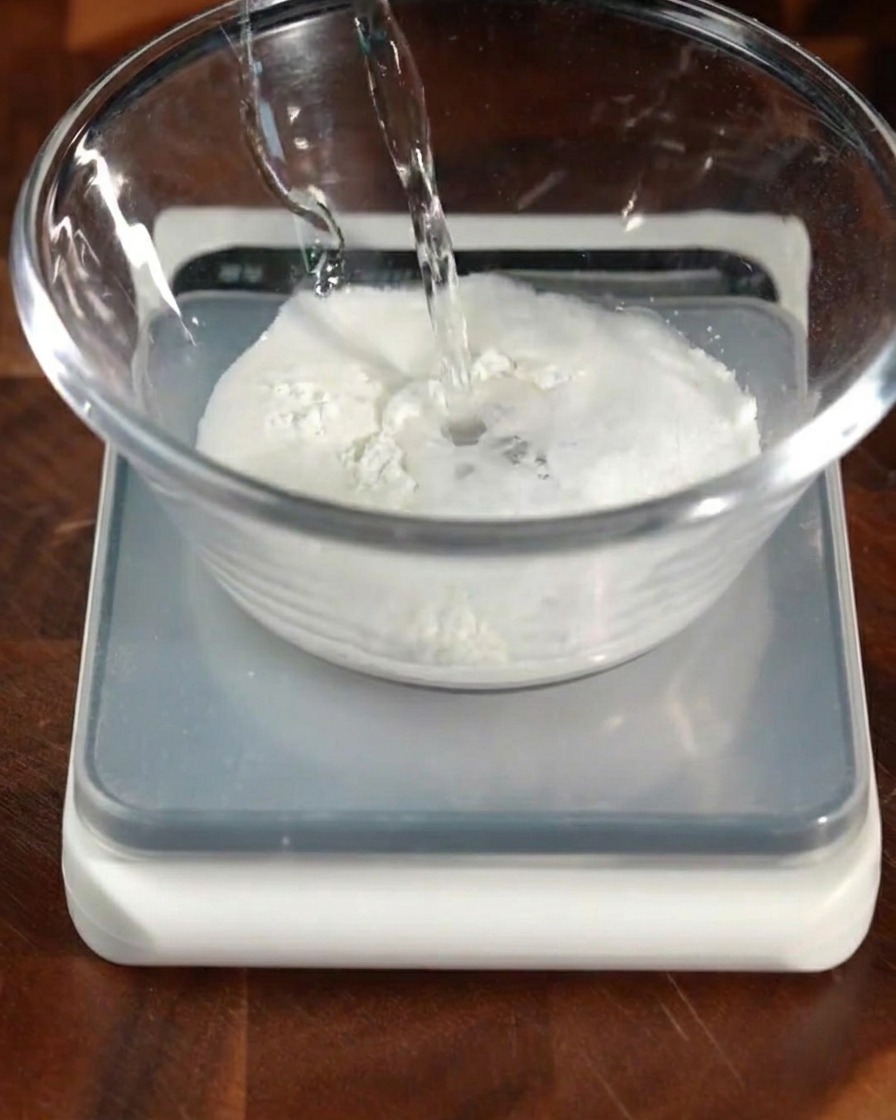 Acqua tiepida con lievito versata in una ciotola contenente farina e altri ingredienti secchi.