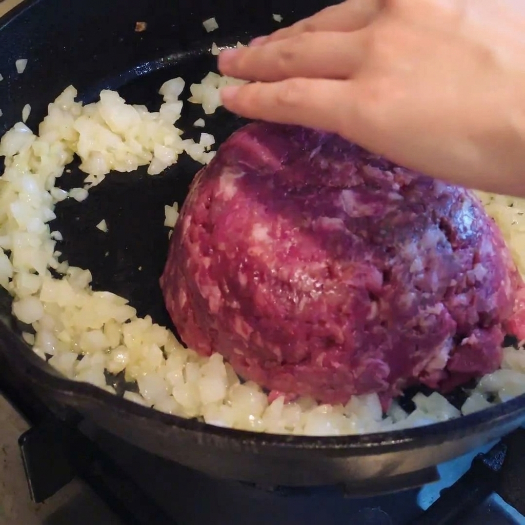 Carne macinata messa in una padella con cipolle precedentemente rosolate, pronta per essere mescolata e condita.