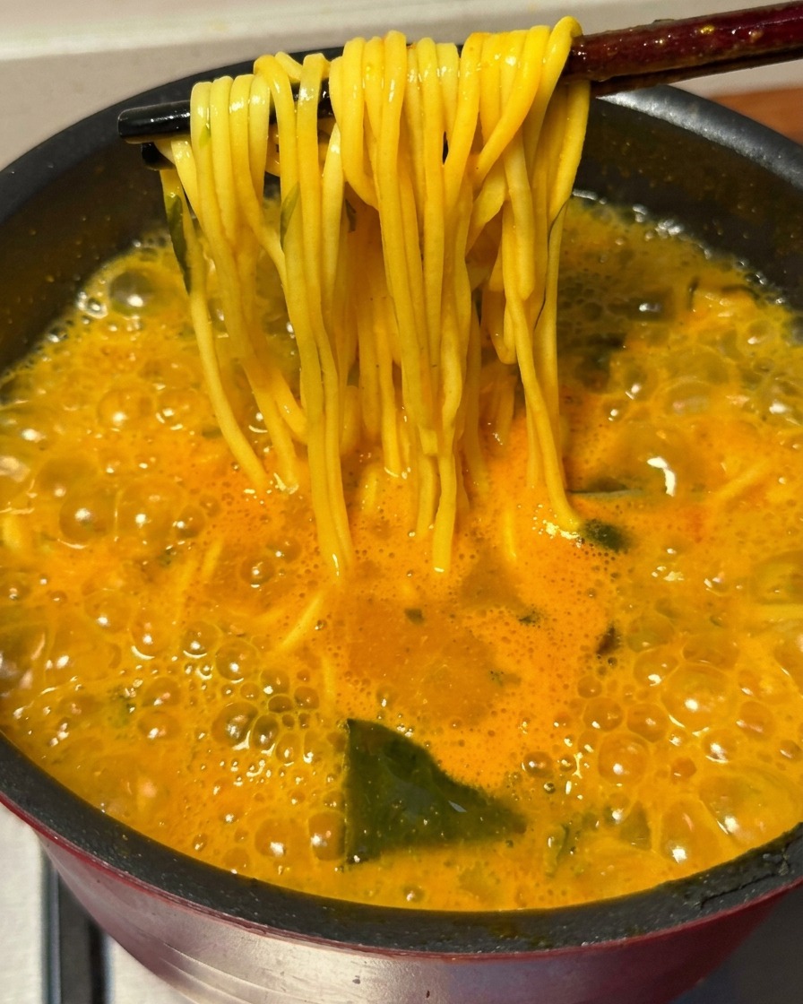 Un paio di bacchette che sollevano un mucchietto di noodles ramen cotti dal brodo piccante che bolle.