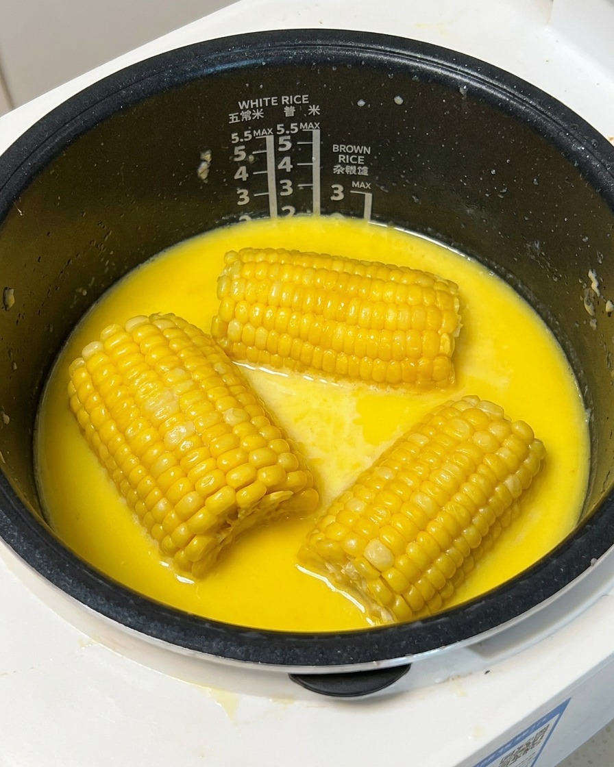 Un pezzo di mais giallo cotto estratto da un brodo lattiginoso nel cuociriso con uno spiedino di legno.