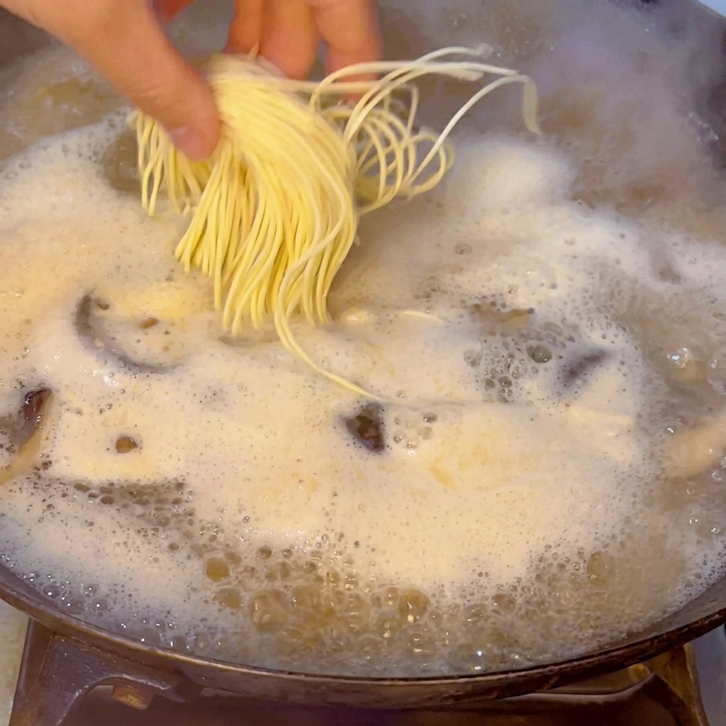 Una mano che fa cadere delicatamente un nido di ramen freschi e gialli nel brodo di pesce bollente.