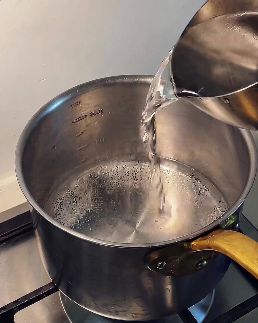 Versare acqua fredda limpida da una caraffa di metallo in una pentola di acciaio inox sul fornello.