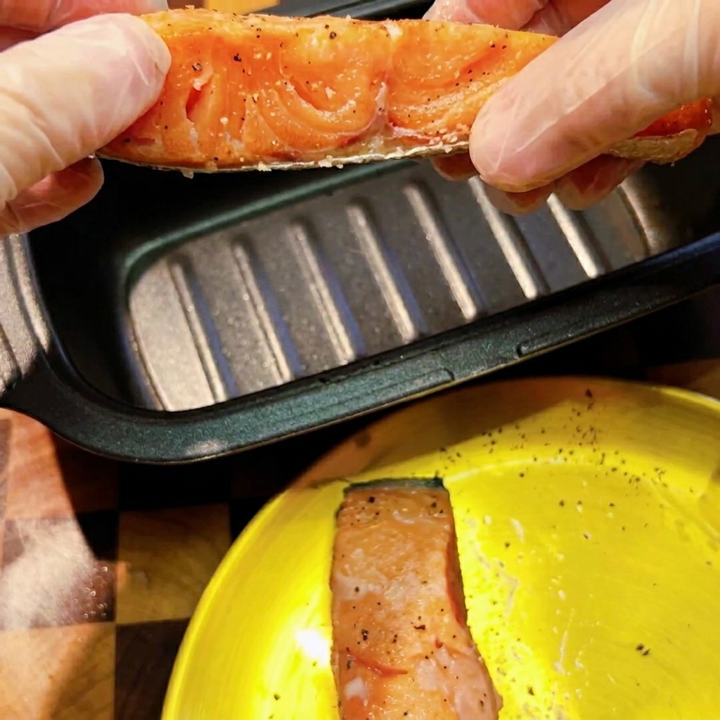Mani con guanti tengono un filetto di salmone crudo condito sopra una padella rettangolare scura per dorare al microonde.