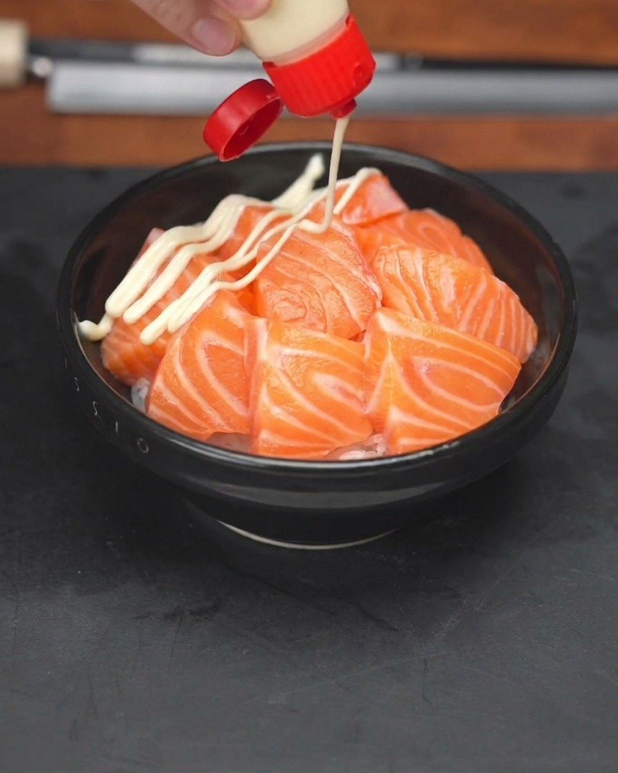 Una mano che spreme una bottiglia di maionese a zig-zag su una ciotola di riso condita con fette di salmone.