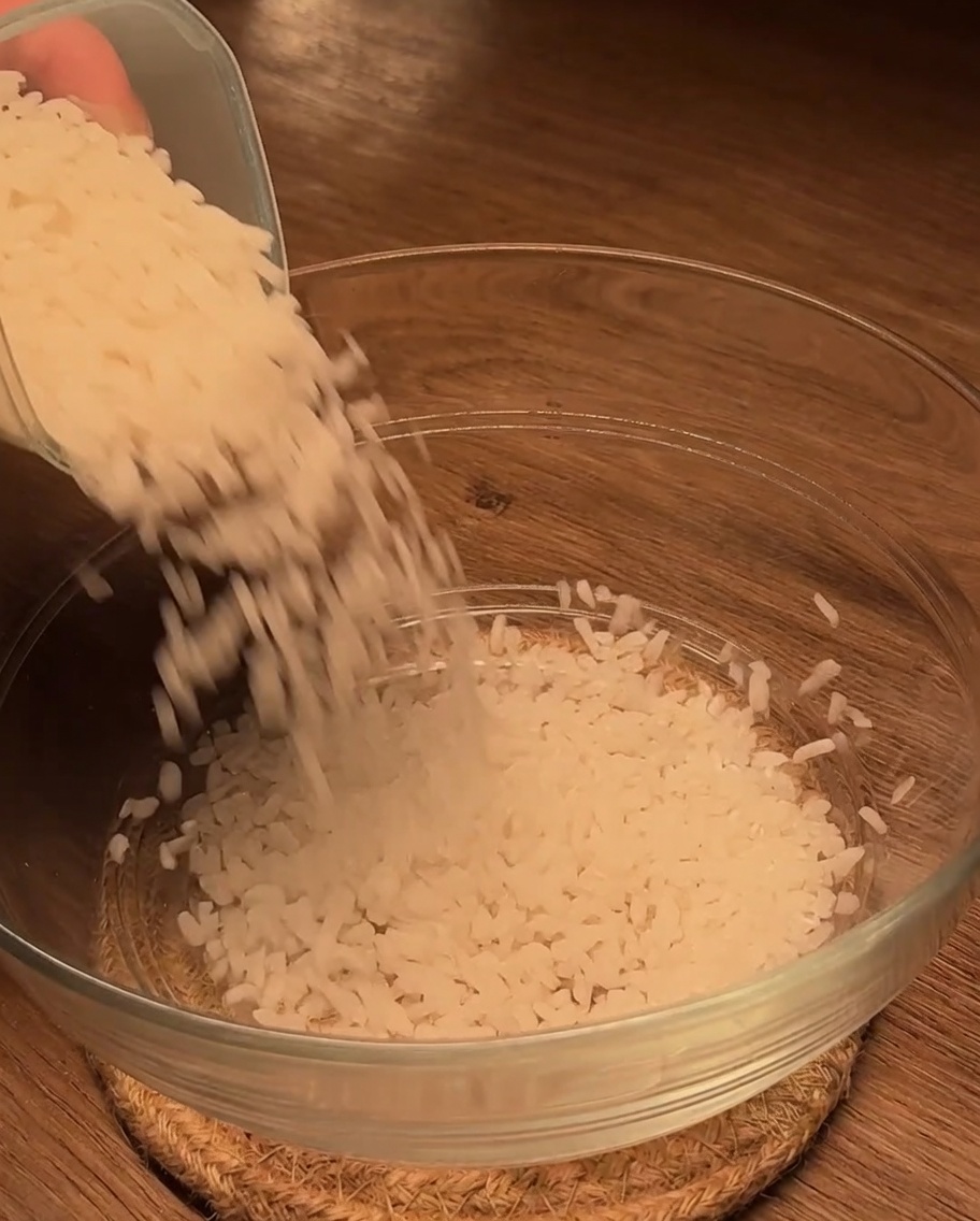 Riso bianco crudo versato in una ciotola di vetro pulita su una superficie di legno.
