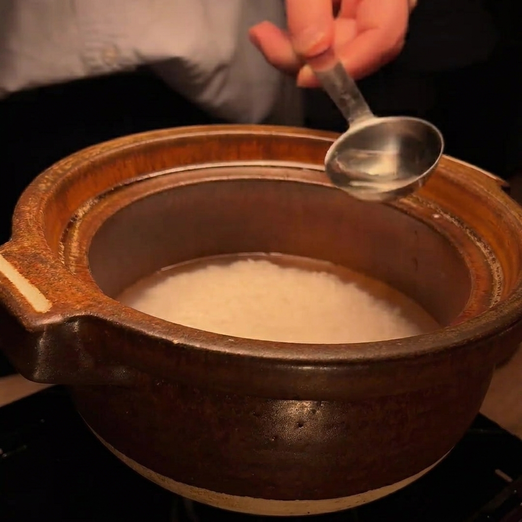 Un cucchiaio di metallo versa una piccola quantità di liquido trasparente, il sake, nel recipiente con riso e acqua.