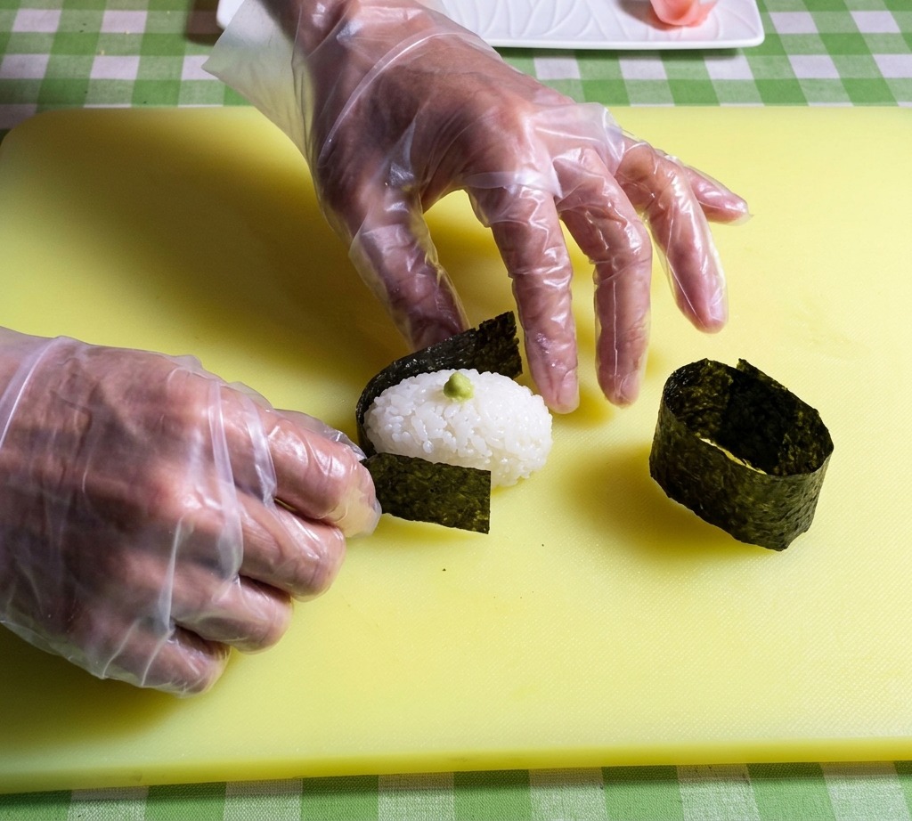 Mani che posizionano una striscia di alga nori scura attorno al bordo di una base ovale di riso per sushi.