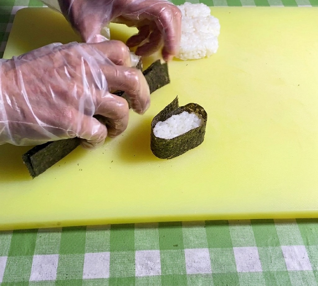Primo piano di mani che premono le estremità di una striscia di nori insieme per fissarla attorno a una base di riso.