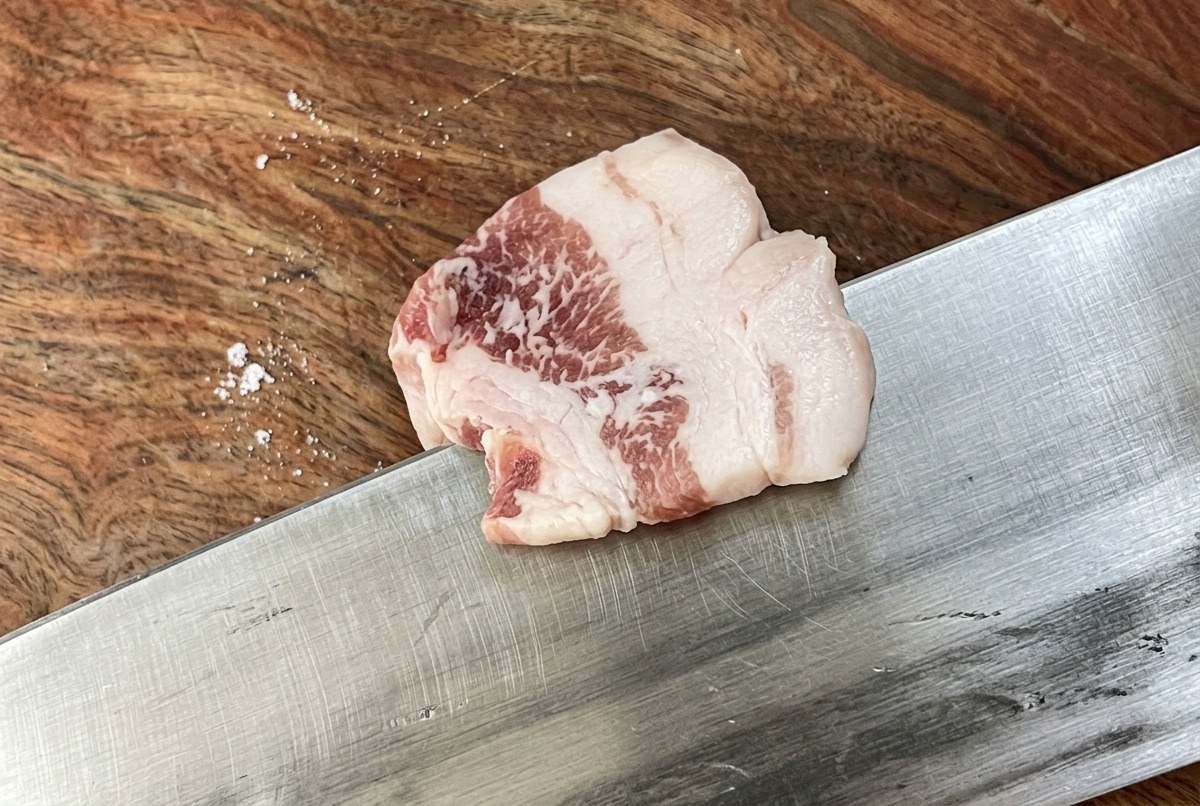 Un coltello affilato che taglia una fetta di pancetta di maiale cruda in strisce sottili e uniformi su un tagliere di legno.