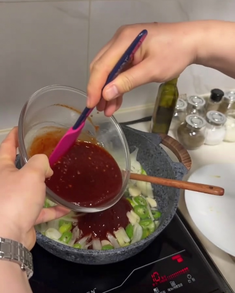 Salsa rosso scuro versata da una ciotola di vetro in una padella contenente verdure saltate.