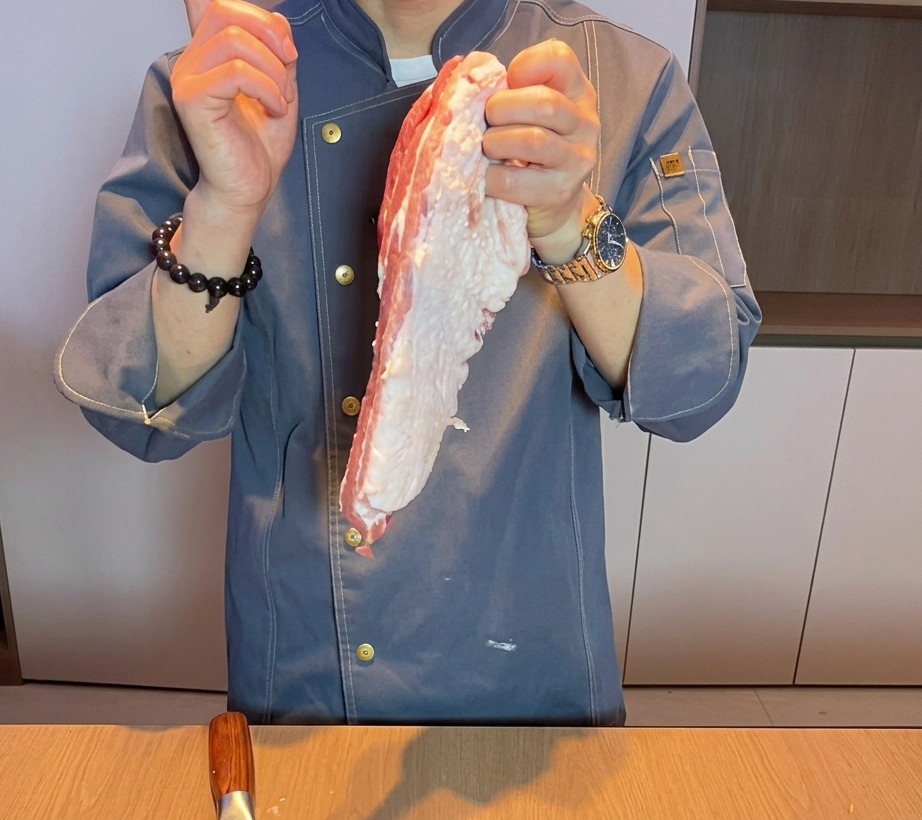 Primo piano delle mani di uno chef che taglia la pancetta di maiale cruda a pezzi spessi su un tagliere di legno.