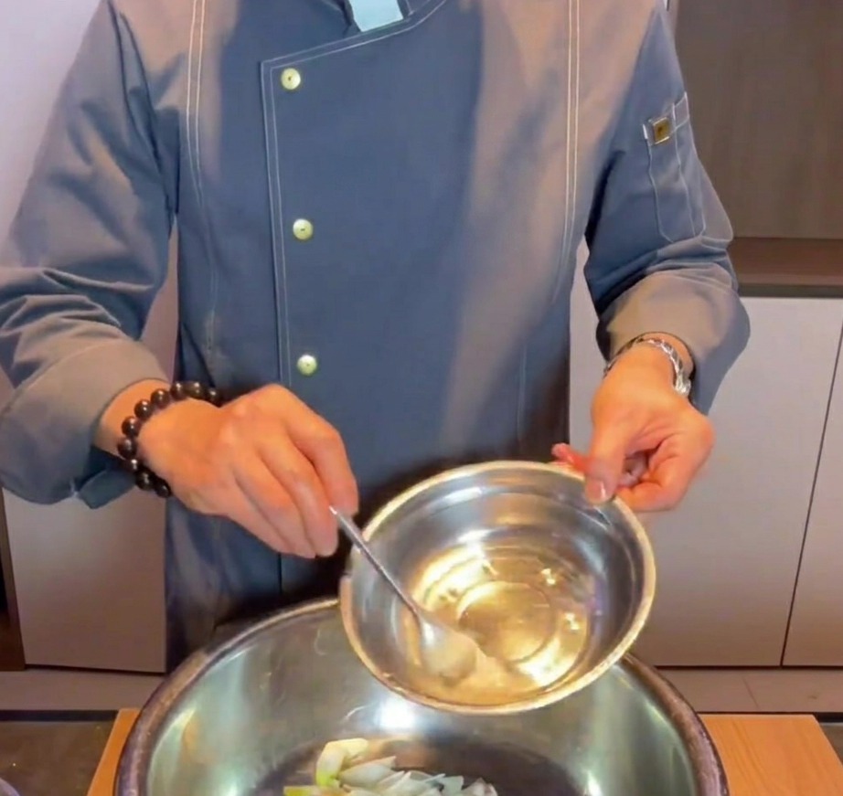 Una mano versa acqua e bicarbonato in una ciotola di metallo con pancetta cruda, zenzero e cipollotto.