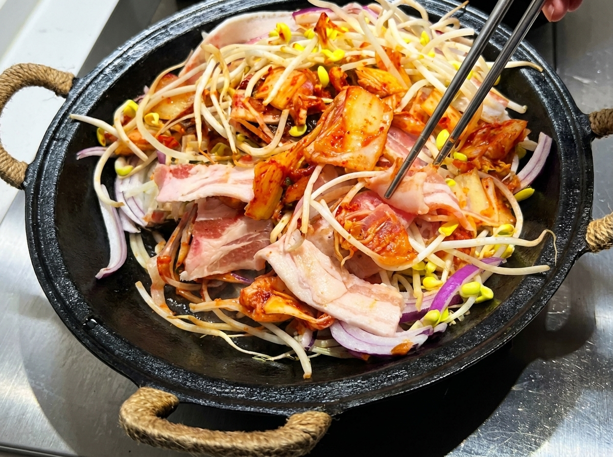 Pinze che mescolano pancetta affettata, germogli di soia, kimchi e salsa insieme in una pentola di ghisa.