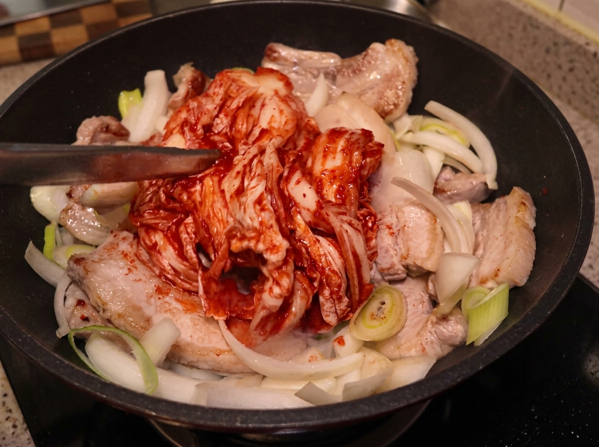 Un grande pezzo intero di kimchi posizionato con le pinze sopra la pancetta e le cipolle.