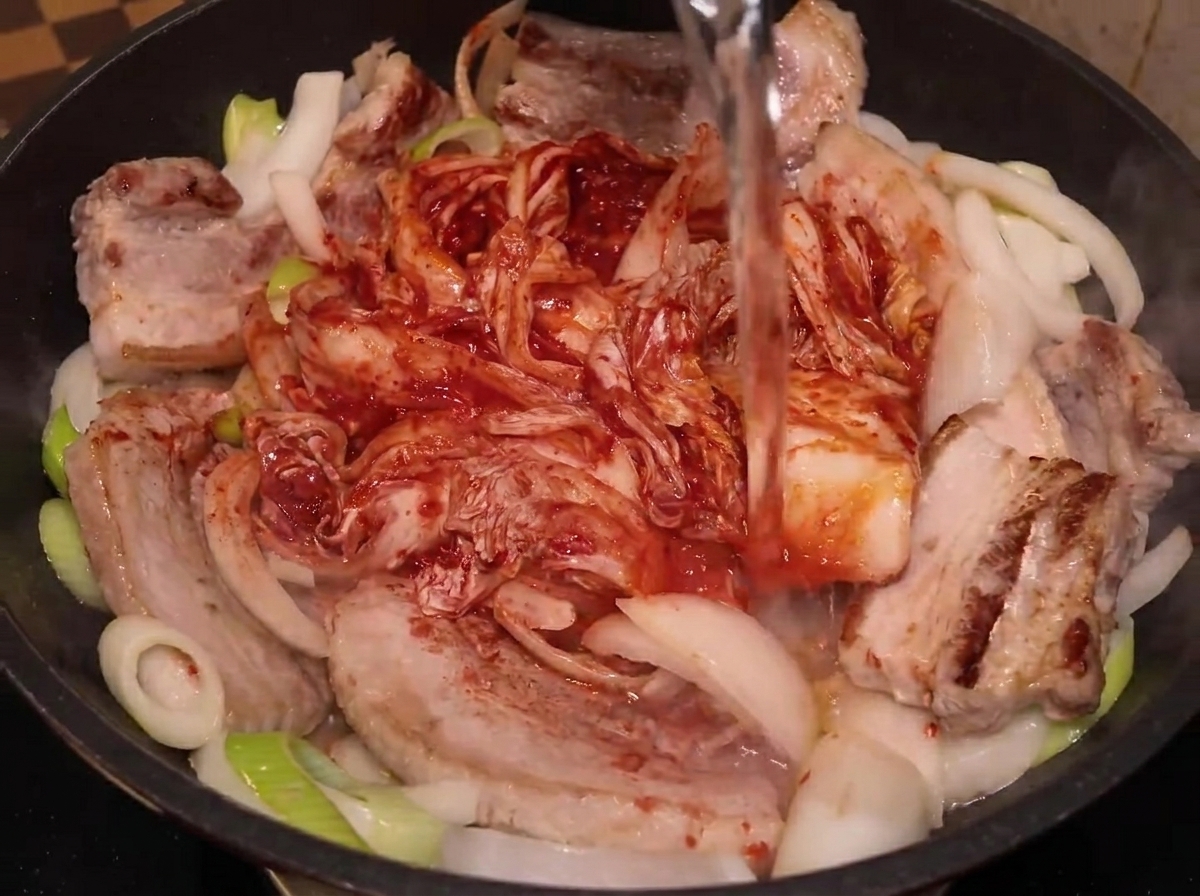 Acqua versata da una bottiglia di plastica in una padella con pancetta, cipolle e kimchi intero.