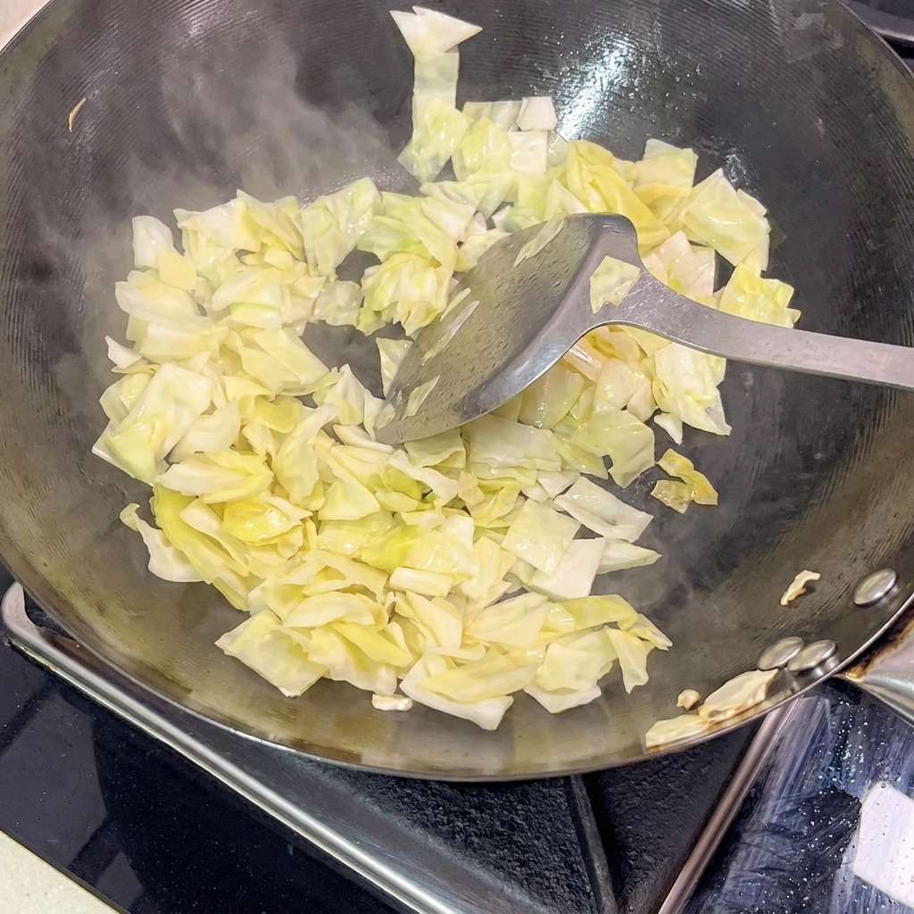 Una spatola di metallo che salta crauti verde chiaro in un wok nero caldo, con vapore visibile che sale dalla padella.