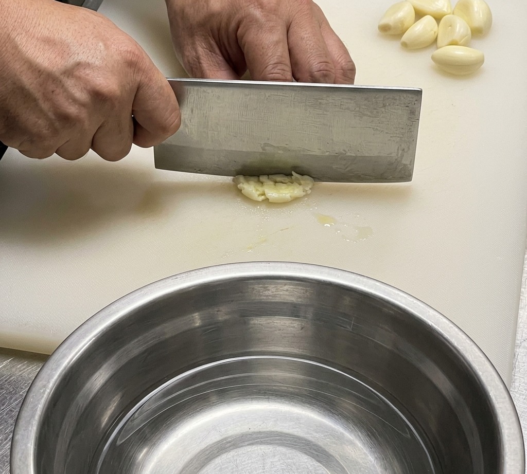 Primo piano di un coltello da chef che trita aglio e zenzero su un tagliere bianco.
