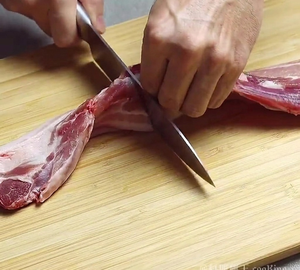 Una persona che usa un coltello da chef per tagliare un pezzo di pancetta di maiale cruda in strisce spesse su un tagliere di legno.