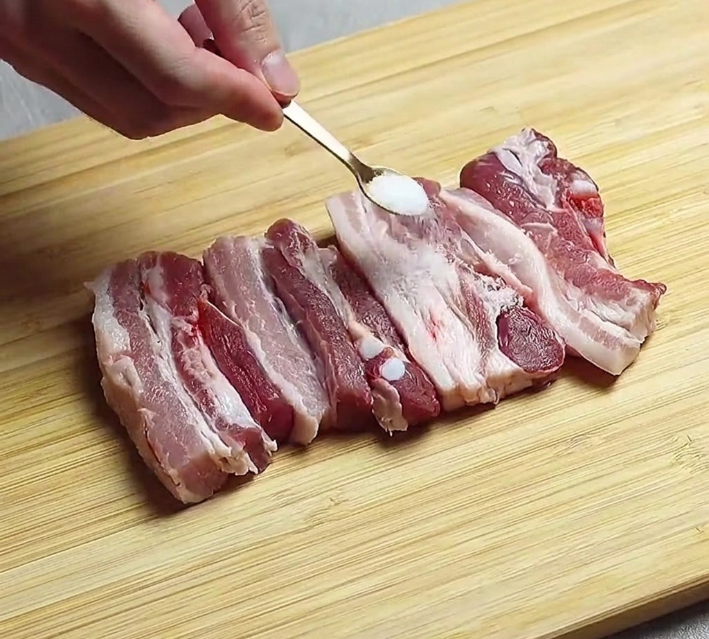 Strisce di pancetta di maiale cruda disposte su un tagliere di legno mentre vengono condite con una spolverata di sale.