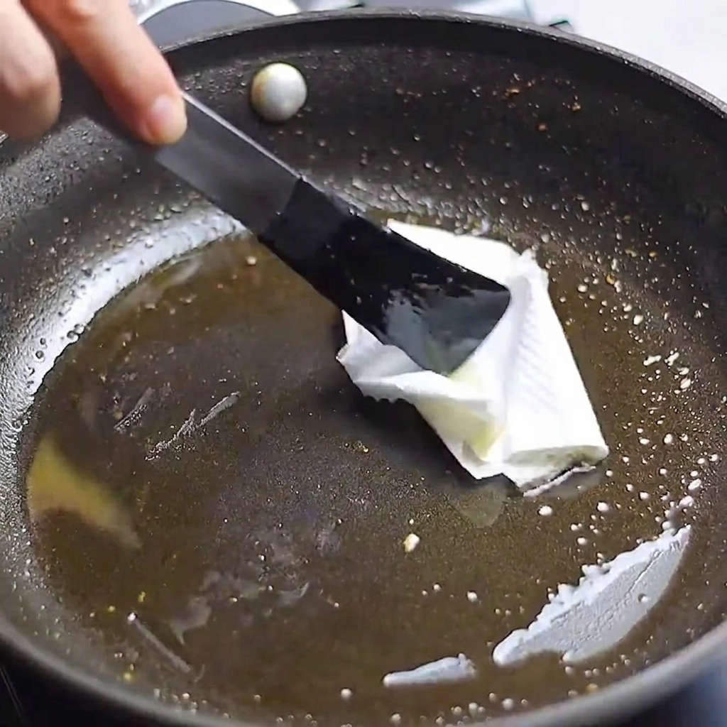Un tovagliolo di carta da cucina tenuto con delle pinze utilizzato per pulire il grasso in eccesso da una padella.