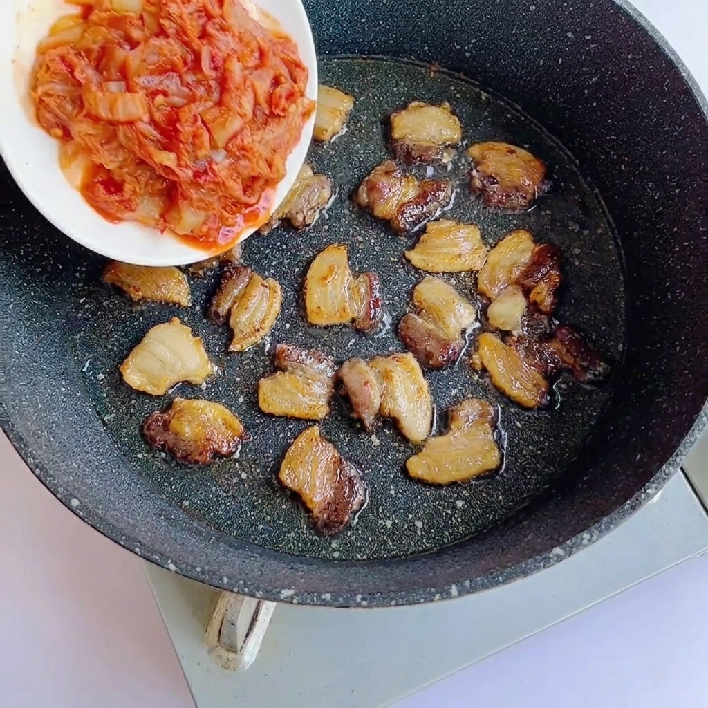 Kimchi tritato aggiunto in una padella con fette di pancetta rosolate.